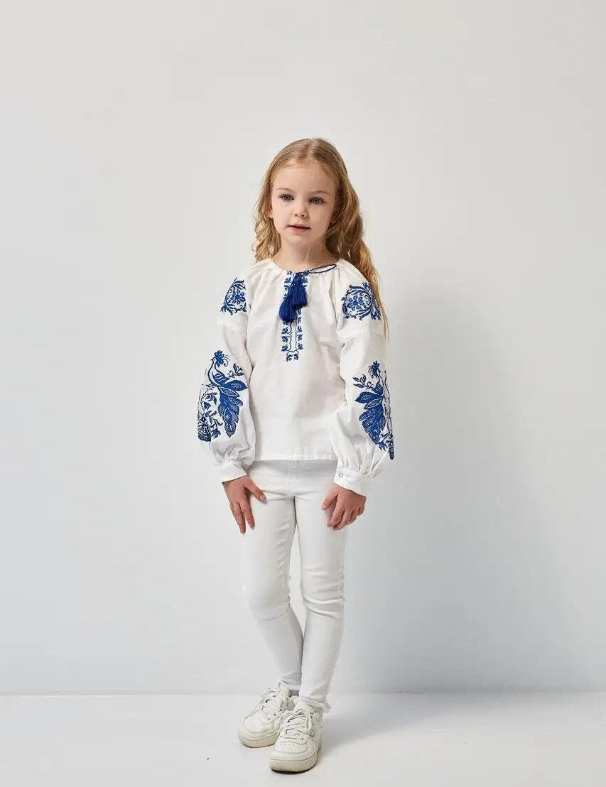 Girl wearing white embroidered Ukrainian shirt with blue phoenix motifs, made from 100% cotton, symbolizing renewal and heritage.
