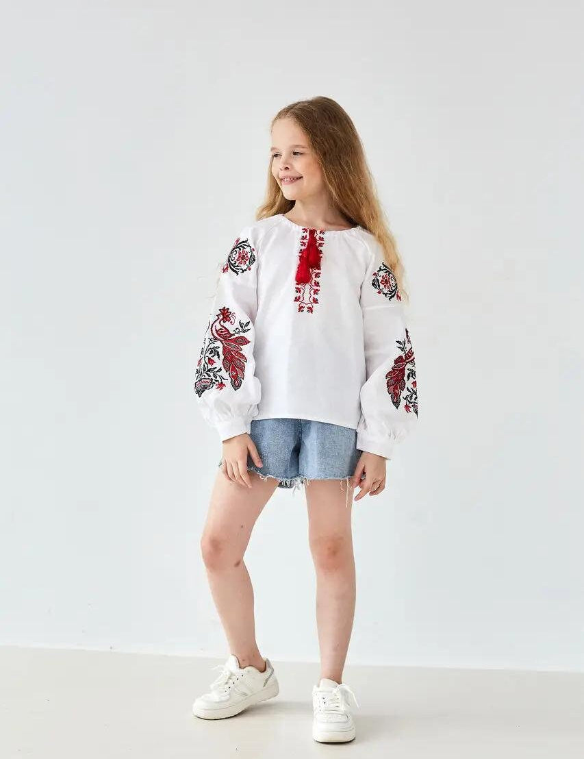 Girl wearing a white Ukrainian embroidered shirt with red and black Slavic motifs, paired with denim shorts, showcasing traditional style.