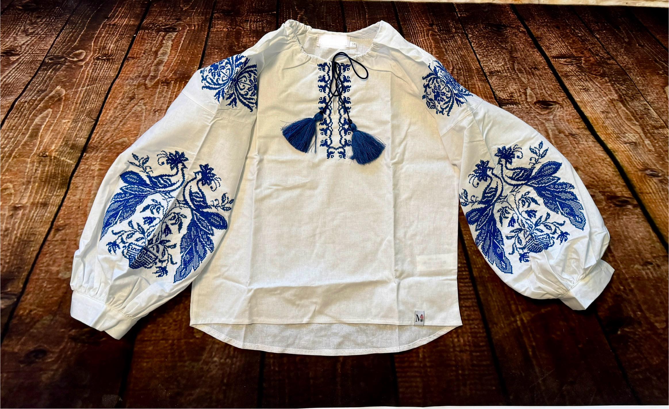 White Ukrainian embroidered shirt with blue phoenix motifs on wooden background, 100% cotton, symbolizes resilience and heritage.