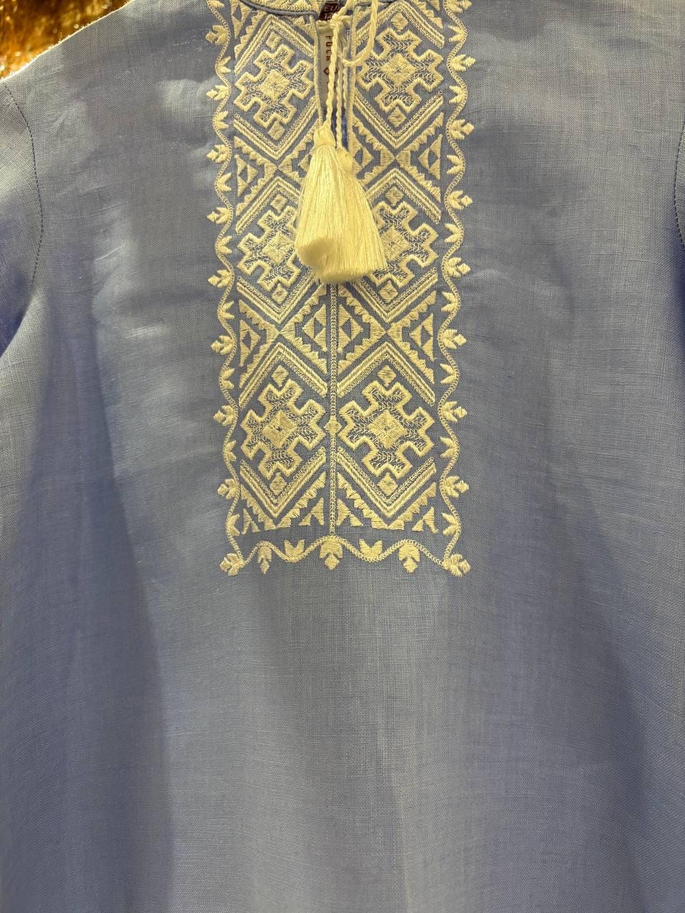 Close-up of a blue Ukrainian Vyshyvanka with intricate white cross-stitch embroidery and tassel detail on natural linen fabric.