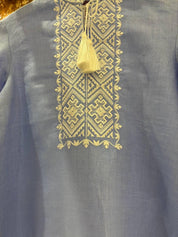 Close-up of a blue Ukrainian Vyshyvanka with intricate white cross-stitch embroidery and tassel detail on natural linen fabric.