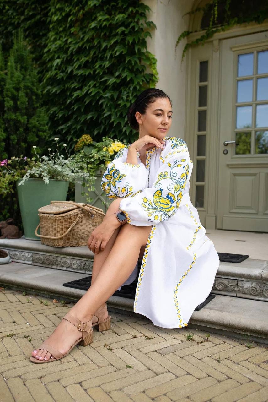 Woman wearing white linen embroidered dress in Ukrainian Vyshyvanka style, sitting on a doorstep, surrounded by garden plants.