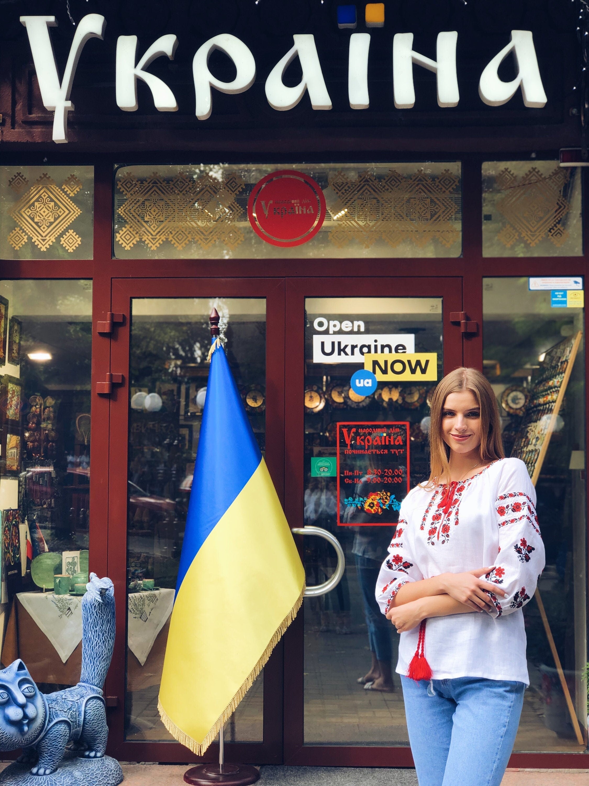 Woman in white vyshyvanka blouse with red embroidery stands in front of a Ukrainian shop with flag.