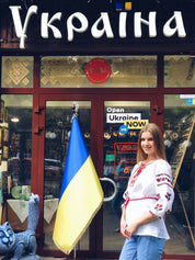 Woman in embroidered Ukrainian vyshyvanka blouse standing outside a store with a Ukrainian flag.