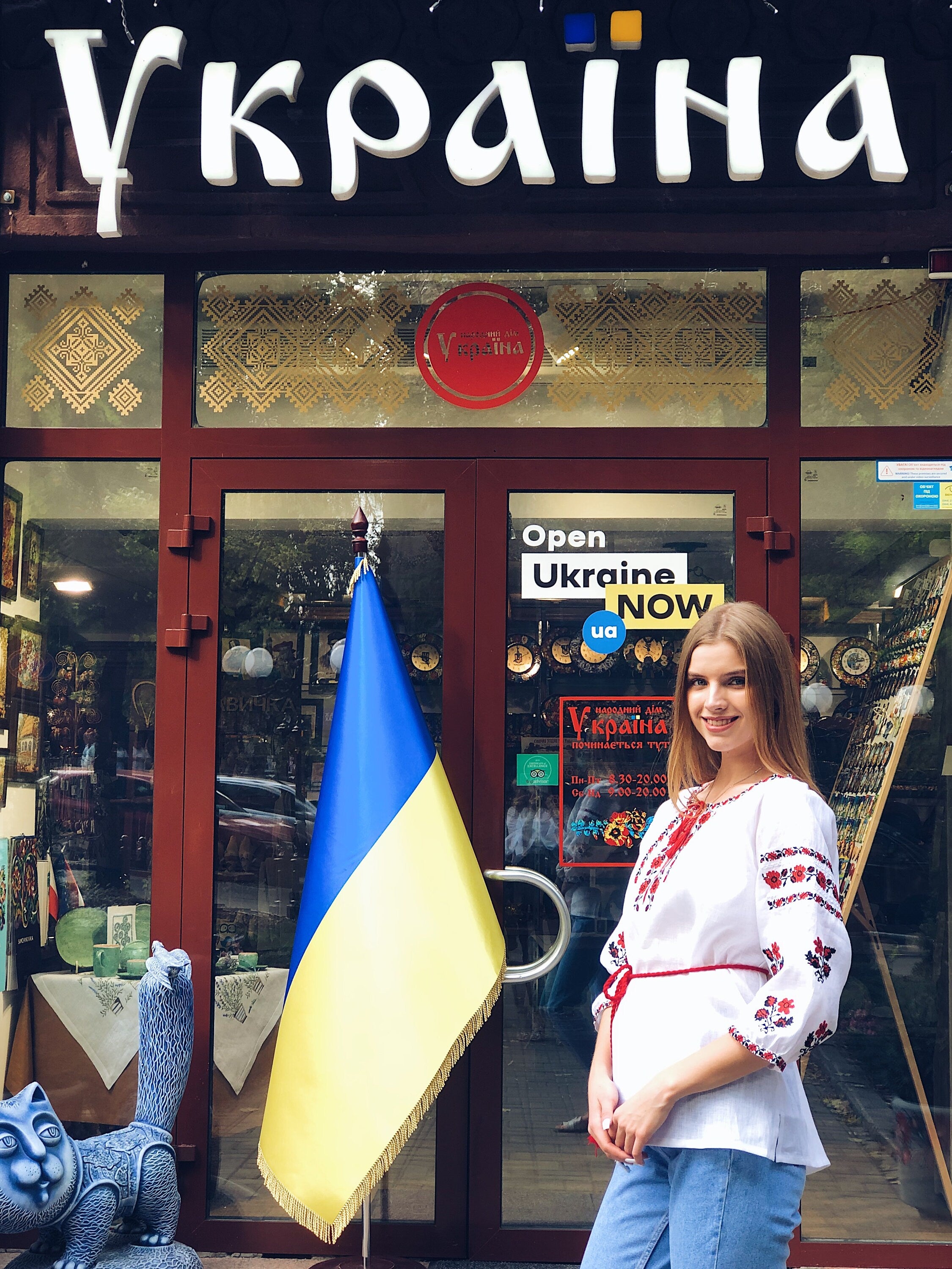Woman in embroidered Ukrainian vyshyvanka blouse standing outside a store with a Ukrainian flag.