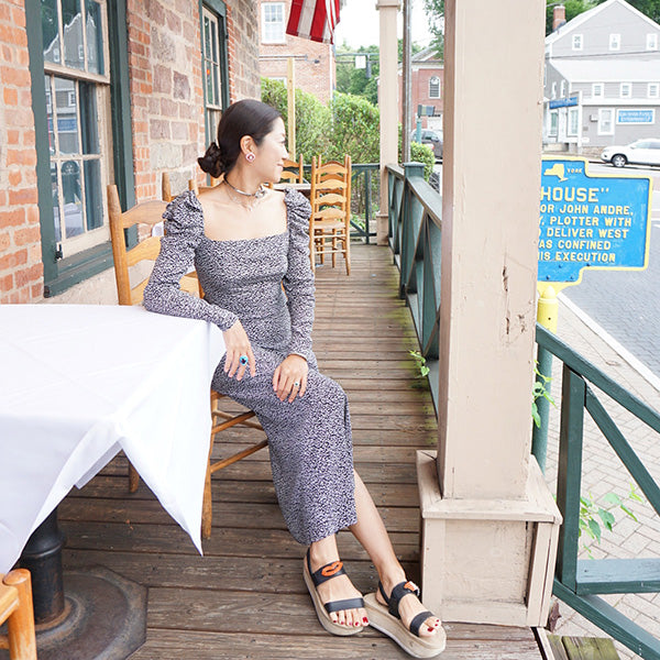 Woman in patterned dress sitting on a porch wearing stylish sandals on a sunny day.