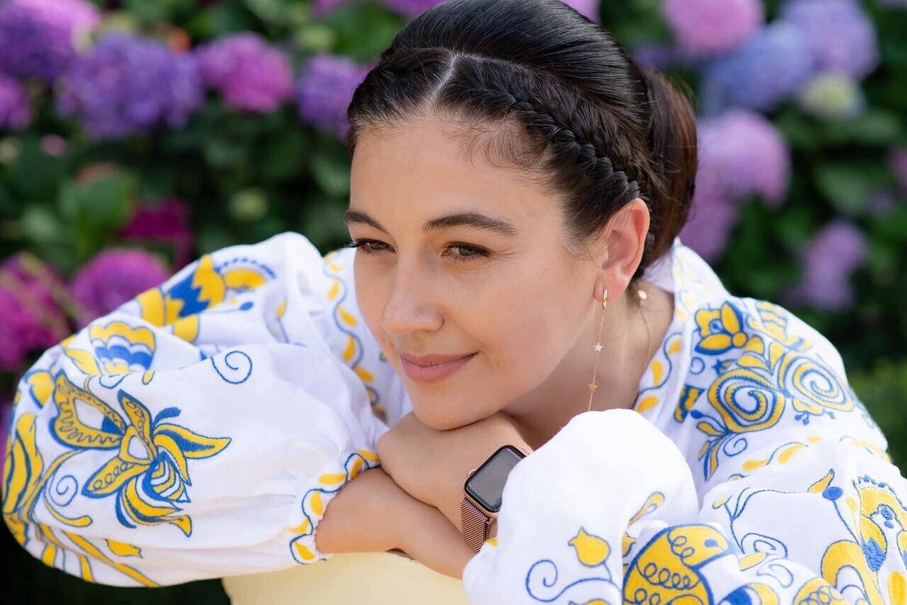 Woman in a white and yellow floral embroidered blouse with braided hair, smiling in a garden setting with vibrant flowers.
