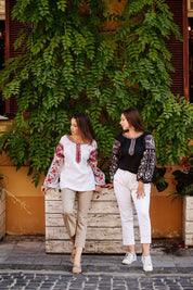 Two women wearing embroidered Ukrainian blouses, one in black and one in white, standing in front of lush green foliage.