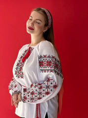 Woman wearing white Ukrainian vyshyvanka blouse with red and black embroidery, standing against a red background.