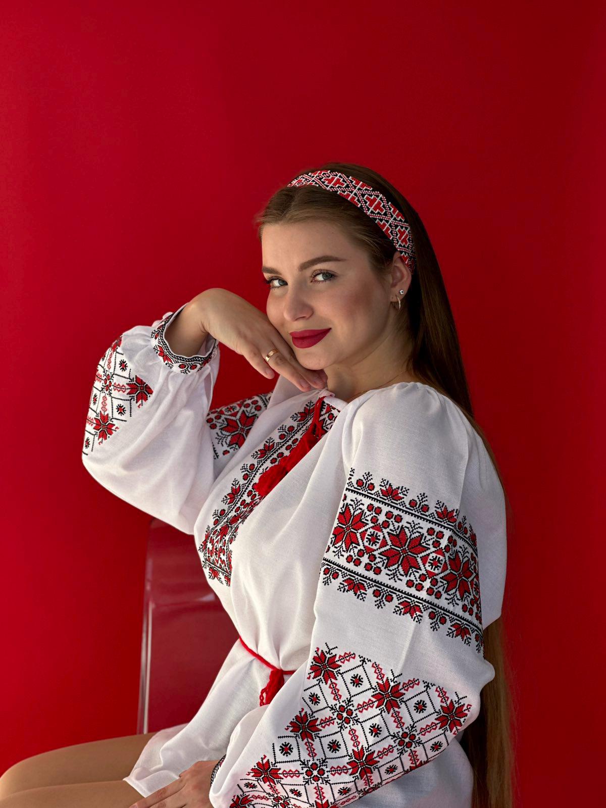 Woman wearing traditional Ukrainian Vyshyvanka blouse with red and black embroidery, sitting against a vibrant red background.