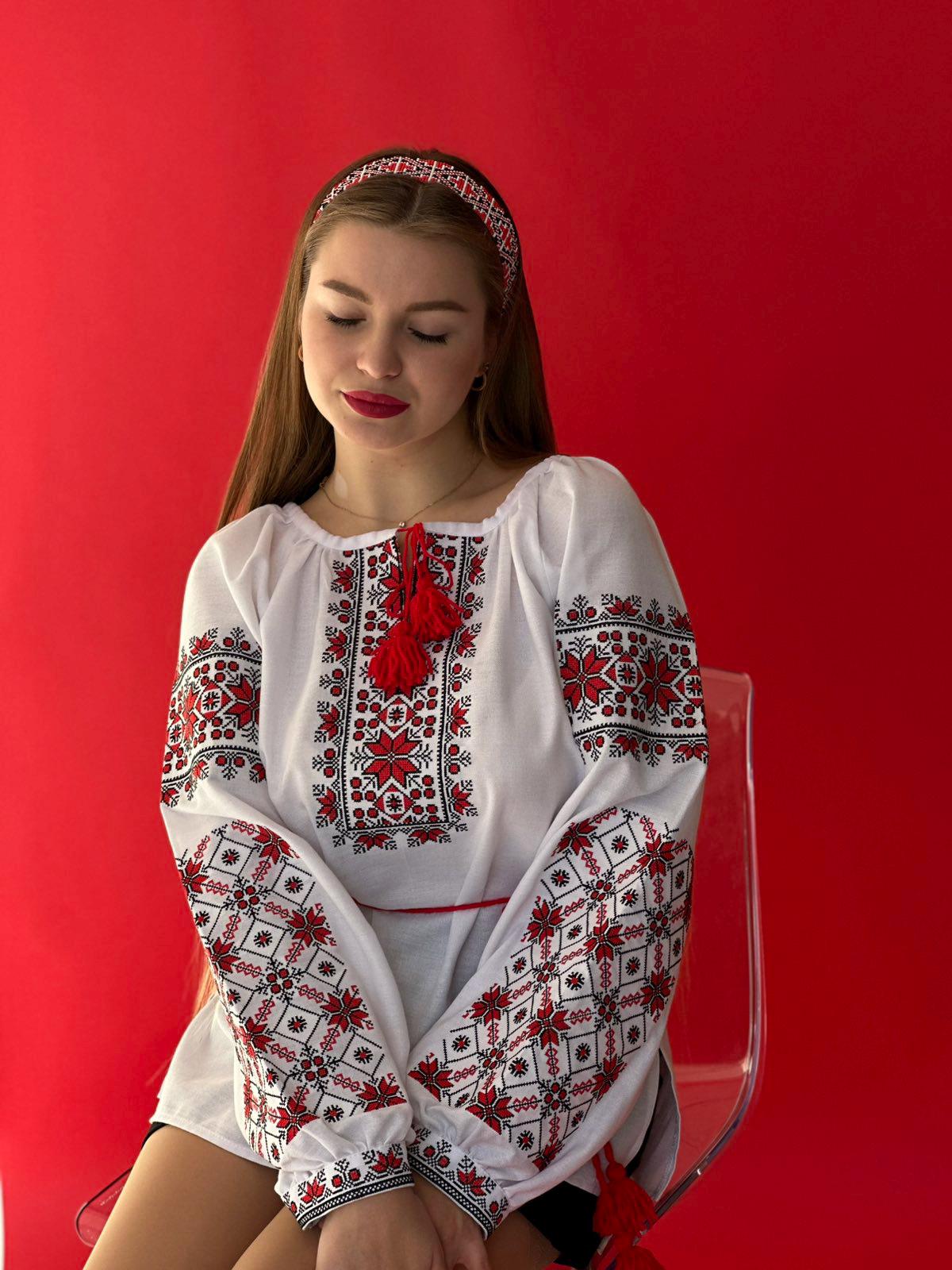 Woman wearing a traditional Ukrainian Vyshyvanka, embroidered blouse on white fabric with red folk patterns, seated against a red background.