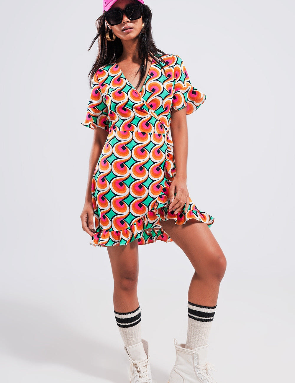 Model wearing Marisol 70s Wrap Ruffle Hem Mini Dress with bold orange geometric print, showcasing V-neckline and frill trim.