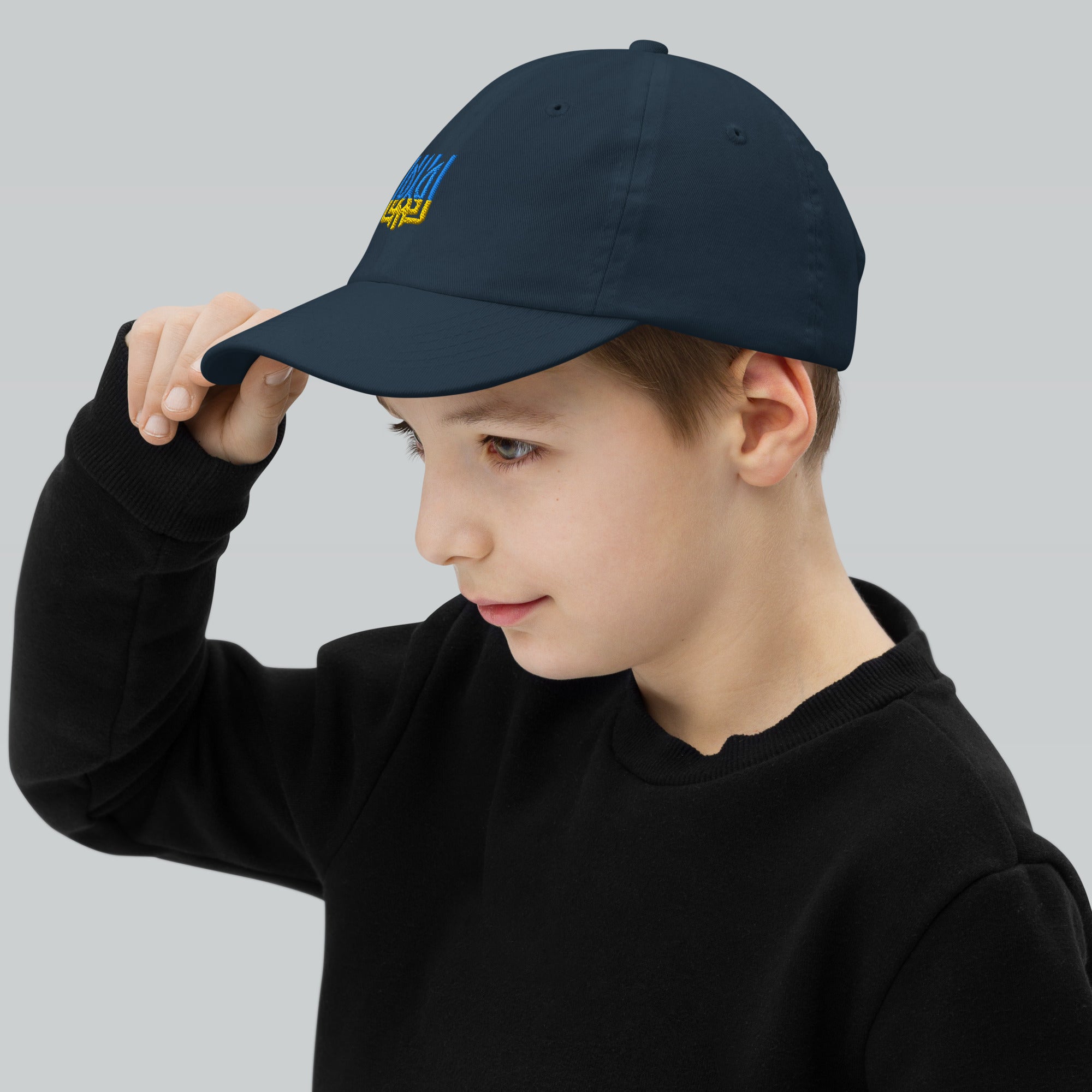 Young boy wearing a youth baseball cap with Ukrainian Tryzub emblem, adjusting the brim. Cap is breathable and adjustable, great for active kids.