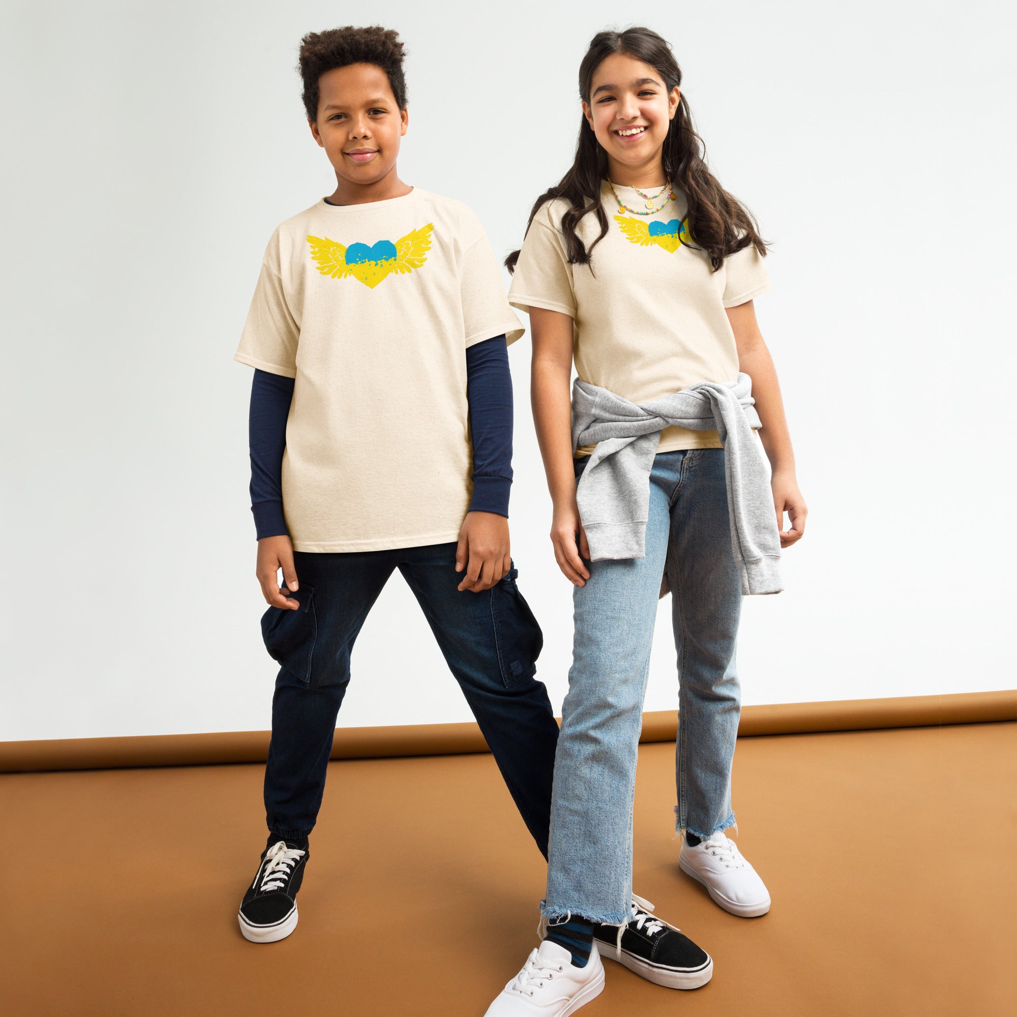 Two kids wearing Ukrainian emblem t-shirts with blue and yellow heart design, standing confidently on a brown background.