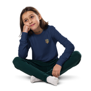 Youth wearing long sleeve shirt with golden Ukrainian Trizub on heart, sitting cross-legged and smiling.